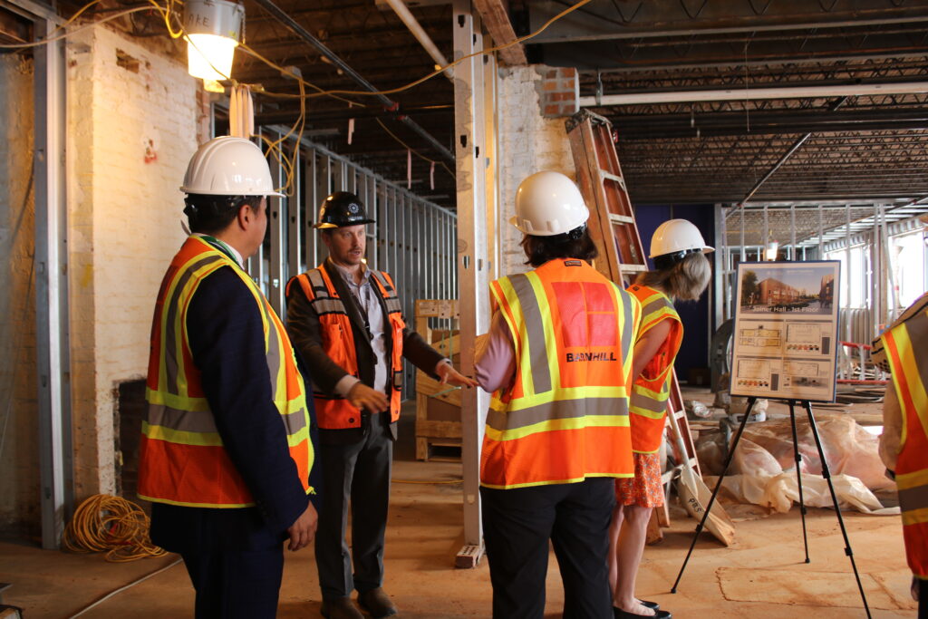 Individuals in safety vests and hard hats examine the interior of Joiner Hall while it was under renovation while others view a stand-mounted rendering of what the completed building will look like. (photo: Beatrice Moss)