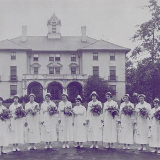 In an old, historic photo, a row of nurses in their uniforms stand holding flowers in front of the three-story main entrance to Watts Hospital, which is now NCSSM-Durham. The architecture is grand and the building is topped with two small chimneys and a cupola.